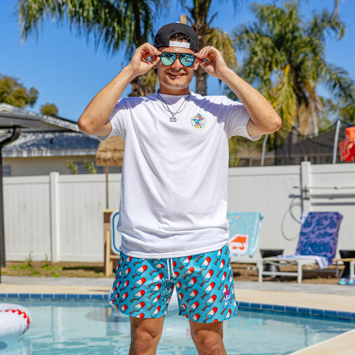 Male model poolside wearing BL101 red, white, and blue popsicle print swim trunks with a white BL101 tee, snapback, and blue mirrored sunglasses.
