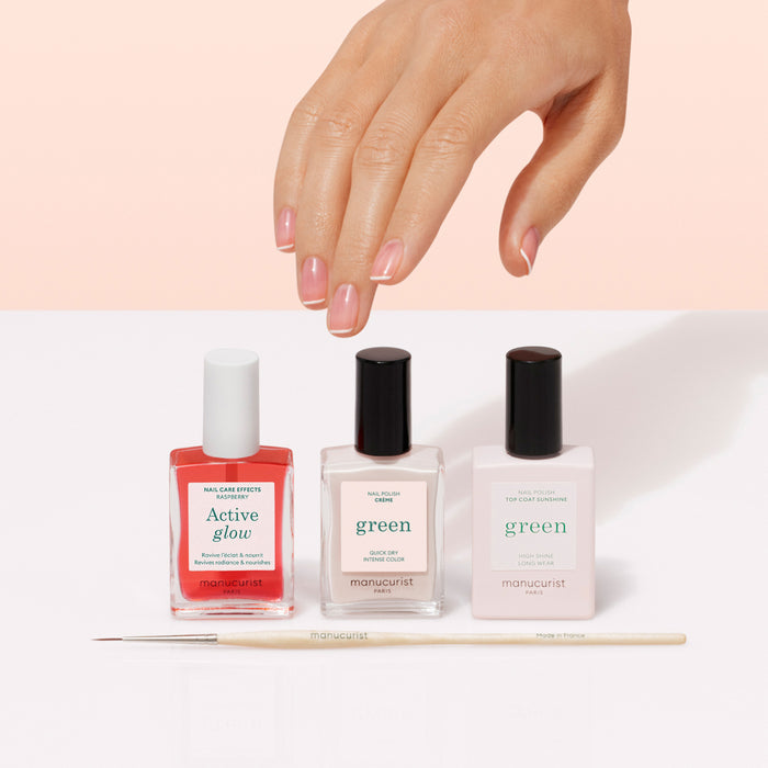 A hand with manicured nails hovers above three bottles of Manucurist French Manicure Glow polish and a thin brush on a white surface, highlighting gel-like shine against a light pink background.