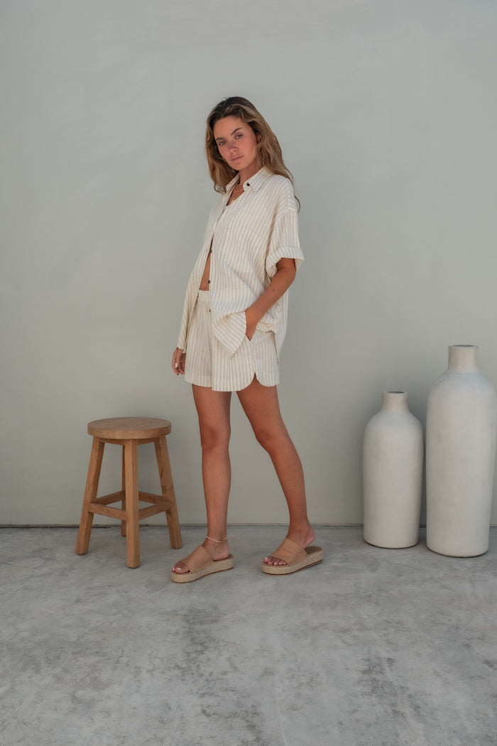 A woman wearing light pink pajama shorts and a matching loose shirt stands against a grey wall with one hand resting in her pockeet looking to the side.
