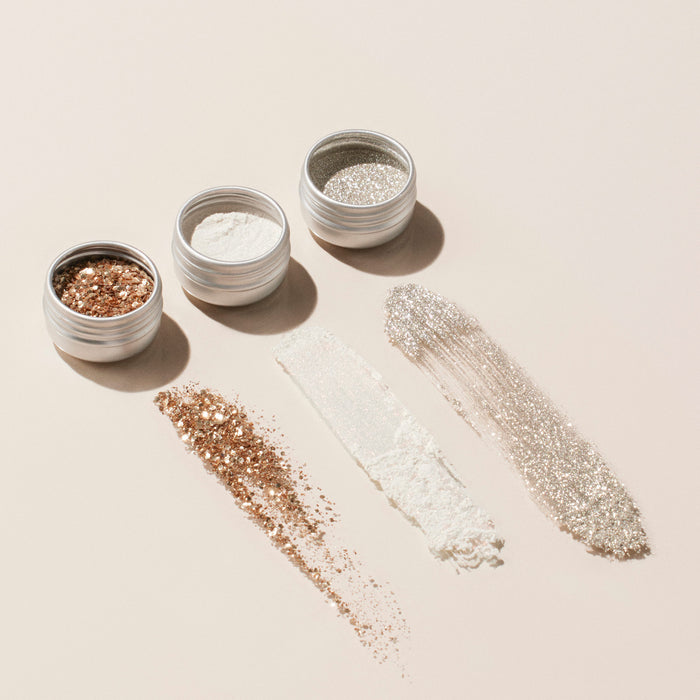 Three small metal tins of Manucurist’s Glitter Trio—Rose Gold, Glazed, and Mirror—are arranged in a row with each powder swatched below its tin. Perfect for adding sparkle to nail polish creations.