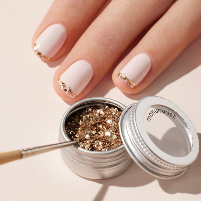 A hand with light pink nails and gold glitter tips rests beside an open jar of Manucurist’s Glitter Trio (Rose Gold, Glazed, Mirror) and a small brush, showcasing the shimmering effect created with these biodegradable powders.