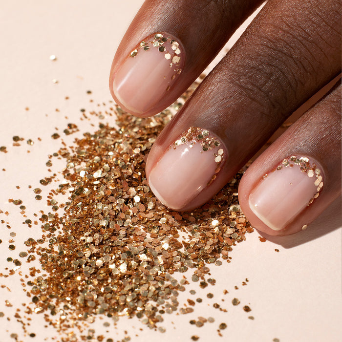 A hand with neatly manicured nails, featuring nude polish and Manucurist’s Glitter Trio—Rose Gold, Glazed, Mirror—on the tips, rests beside a pile of biodegradable glitter on a light beige surface.