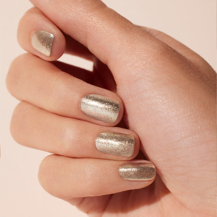 A hand with manicured nails painted in Manucurist's Glitter Trio—Rose Gold, Glazed, and Mirror—sparkles with biodegradable glitter against a soft neutral background.