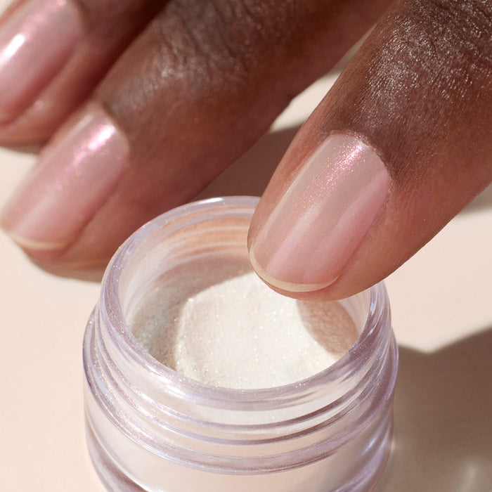 A close-up of a hand with shimmery pink nails dips a finger into a small jar of Glitter Trio - Rose Gold by Manucurist, a fine white glitter powder perfect for nail art or makeup.