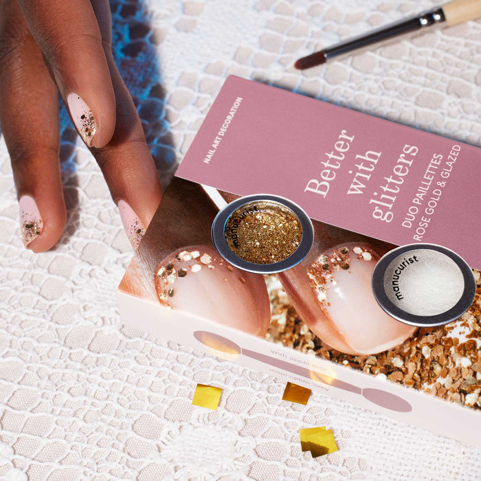 A hand with nude nails, adorned with Manucurist’s Glitters Duo Rose Gold  Glazed, rests on lace beside a pink and white "Better with glitters" box, showcasing rose gold and pearl powders for a stunning pearlescent shine.
