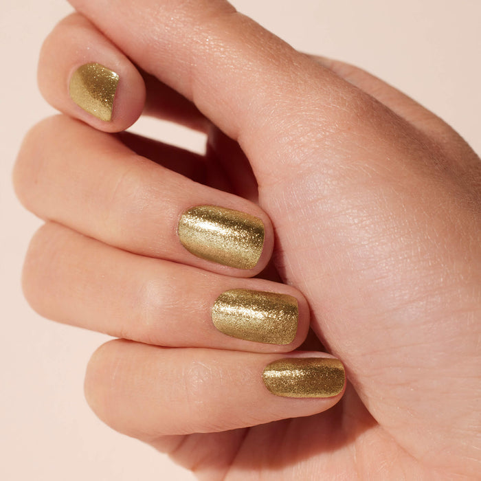 A close-up of a hand with short nails coated in Manucurist Golden Chrome Powder, creating a sparkly gold effect against a neutral background.