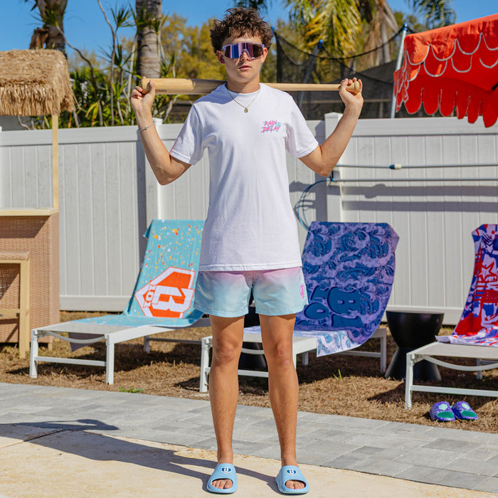 Male model poolside wearing BL101 pink to white ombre swim trunks, white Rain Delay tee, and light blue slides, resting a bat across his shoulders.