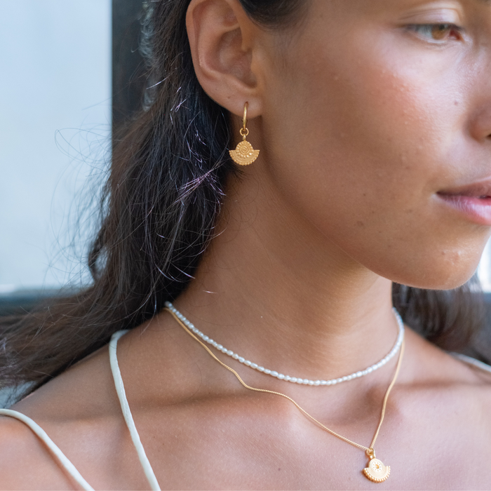A close- of a woman wearing gold sunburst eauprrings, a gold necklace with a sunburst pendant, and a pearl necklace.