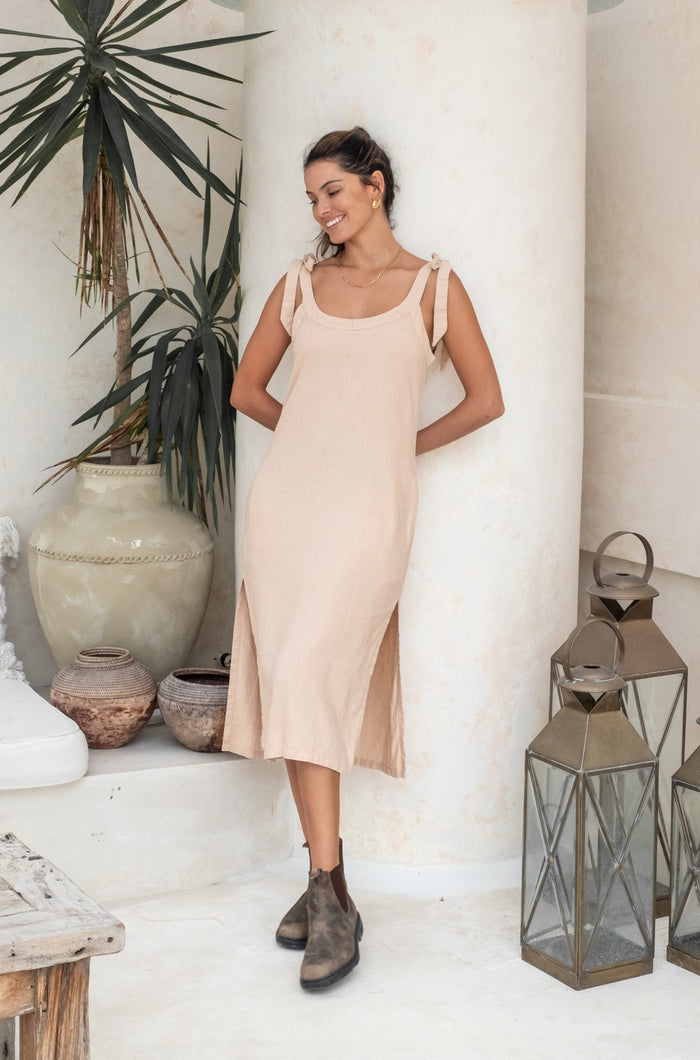 A woman in a light beige midi dress with tied straps, brown Chelsea boots, and holding a woven basket with flowers, stands in a minimalist room.