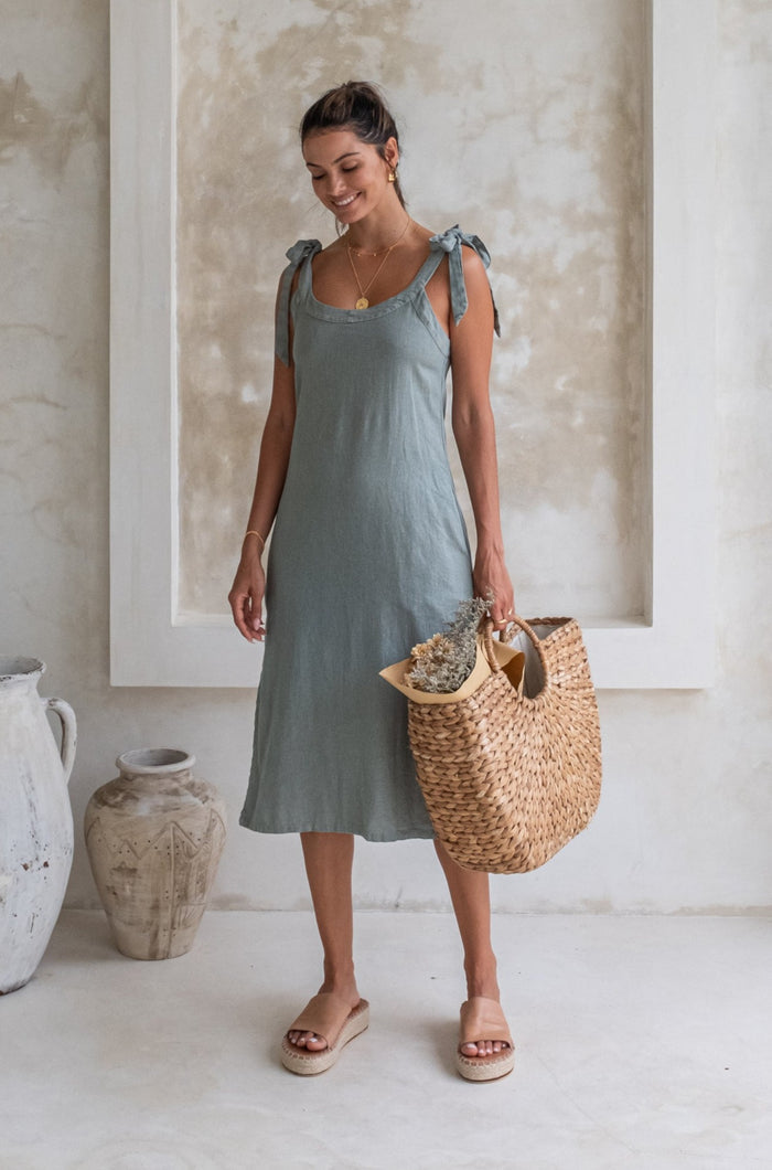 A woman in a light green midi dress with tied straps and beige espadrille sandals stands in a minimalist room, holding a woven basket with flowers and smiling.