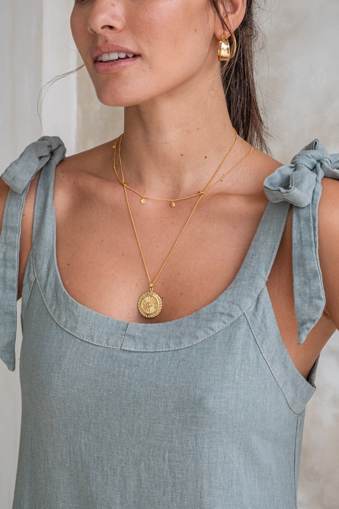 Close-up of a woman wearing a light green dress with tied straps, gold earrings, and layered gold necklaces.