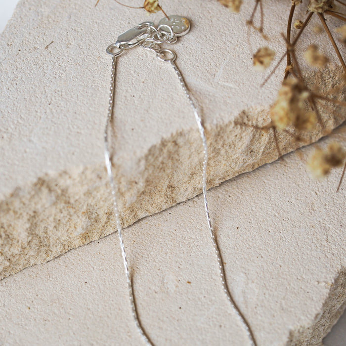 A delicate silver anklet, displayed on beige stone with dried flowers.