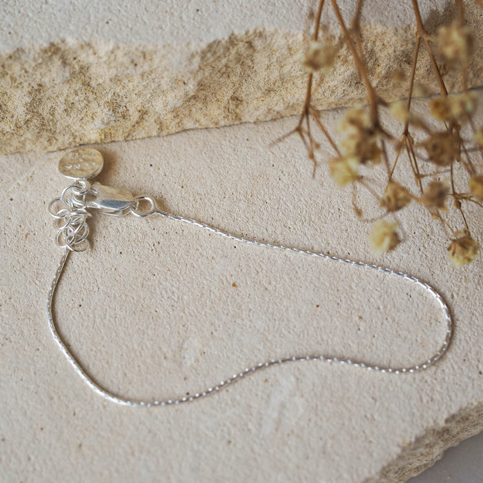 A delicate silver chain bracelet, displayed on beige stone with dried flowers.