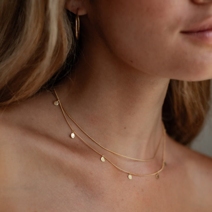 Close-up of a woman's neck wearing a thin gold chain necklace and gold hoop earring.