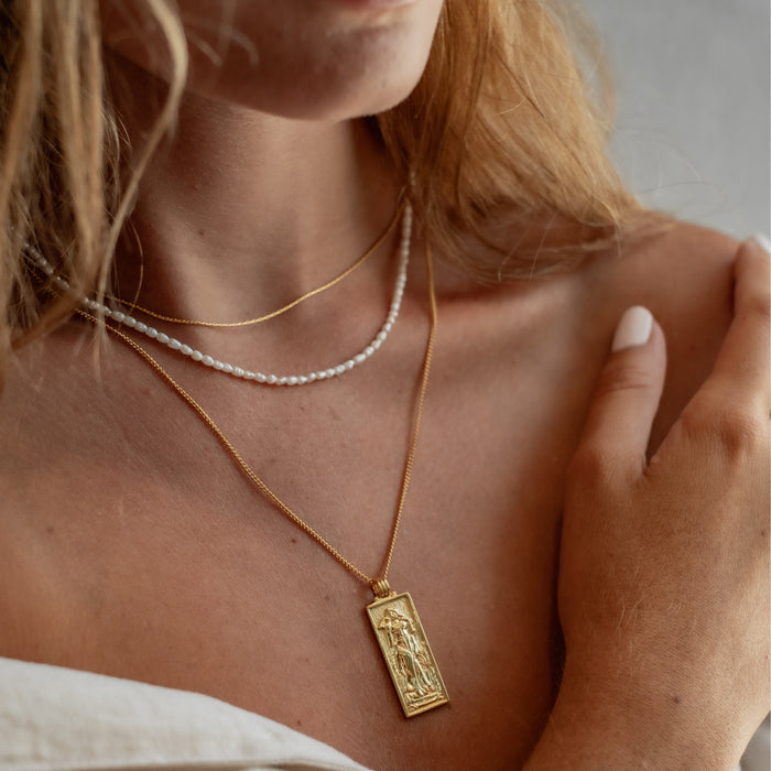 Close-up of a woman's neck and shoulders wearing layered necklaces, including a gold necklace with a rectangular pendant and a pearl necklace.
