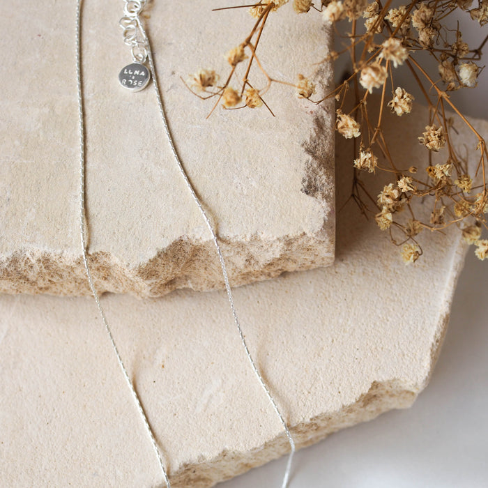 A delicate silver chain bracelet, displayed on beige stone with dried flowers.