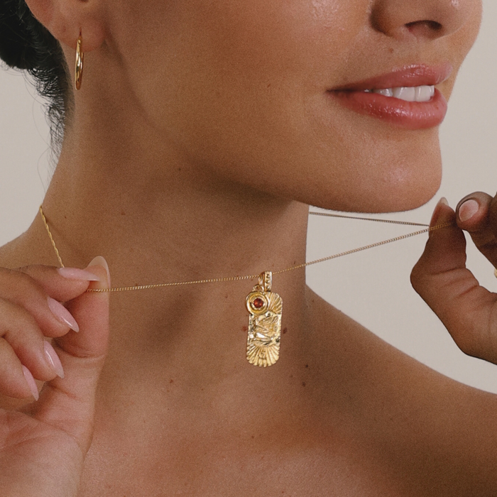 Close-up of a woman wearing a gold necklace with a pendant, against a neutral background.