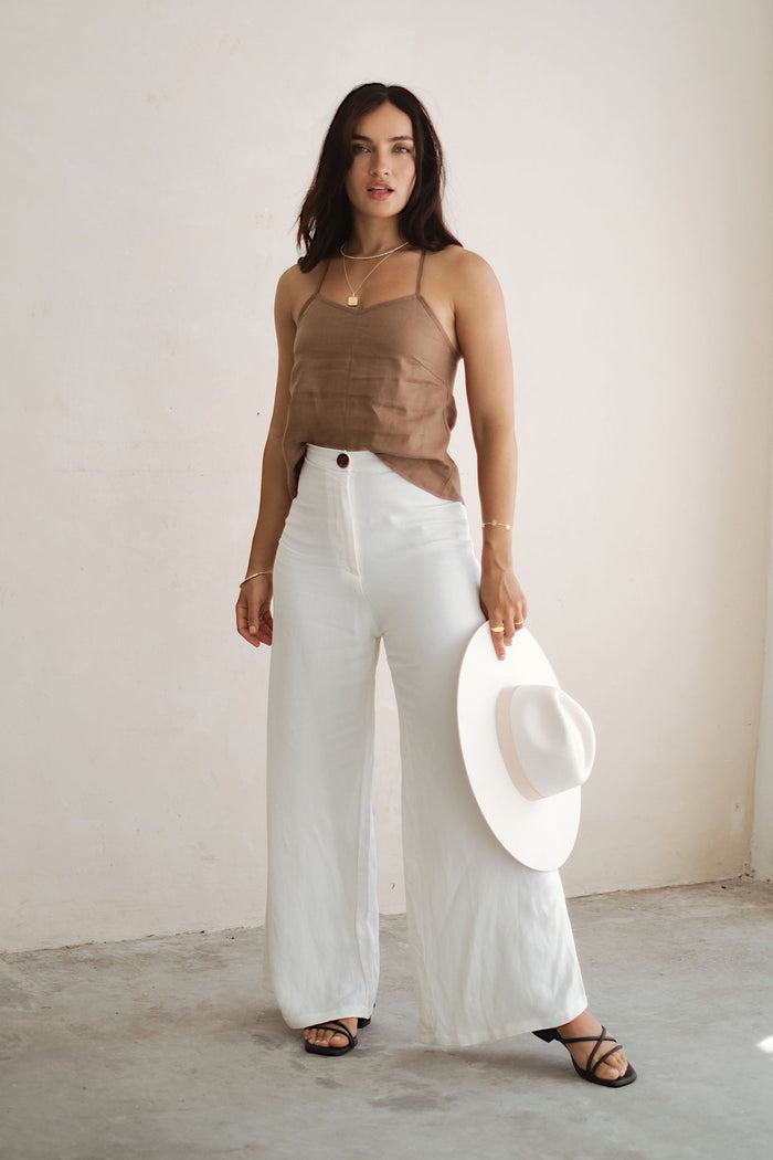 Woman wearing a brown top and white pants holding a hat indoors.