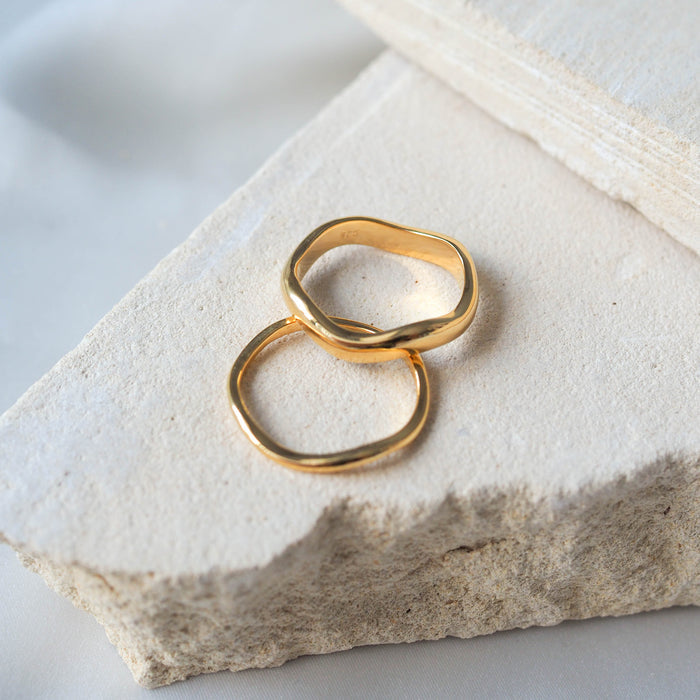 A pair of gold rings with organic, wavy designs displayed on beige stone