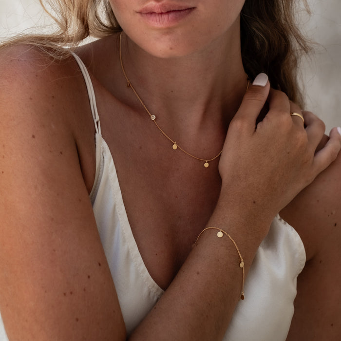 A woman wearing a matching gold necklace and bracelet with small circular charms, shown close-up from the chest up.