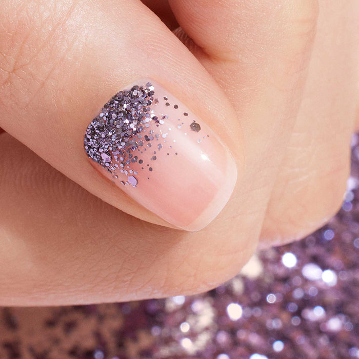 A close-up of a fingernail with clear base polish, decorated at the cuticle with Manucurist's Lilac Biodegradable Glitter that fades toward the tip. The background is accented with scattered silver glitter for a modern nail art look.