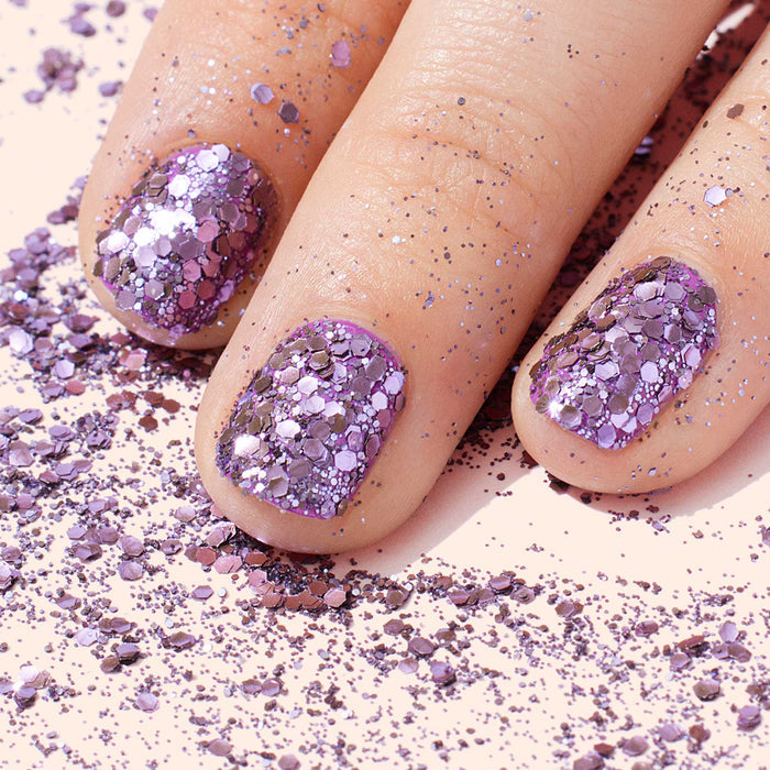Close-up of three fingers with nails painted in sparkling mauve polish and elegant nail art, surrounded by Manucurist Lilac Biodegradable Glitter scattered on a pale pink background.
