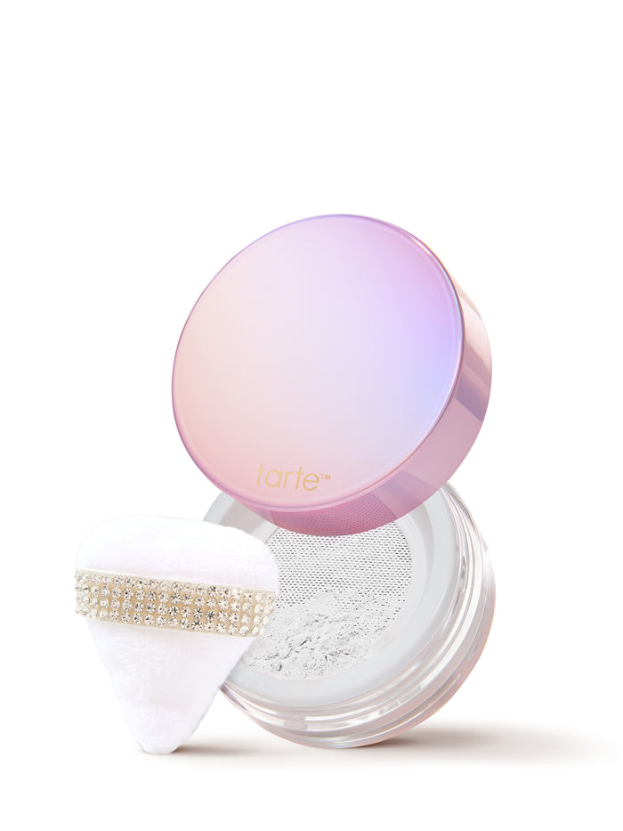 Tarte iridescent round compact open to reveal pressed powder, soft white puff resting nearby; both positioned beside a striped pastel decorative box on a clean white background.
