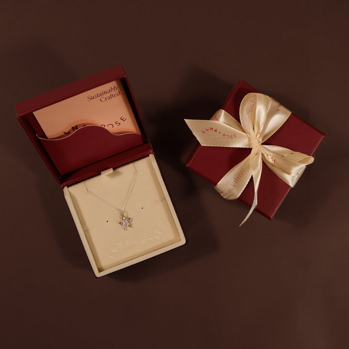 A silver necklace with a star-shaped pendant displayed in an open cream jewelry box next to a closed dark red gift box tied with a cream satin ribbon, on a dark brown background.