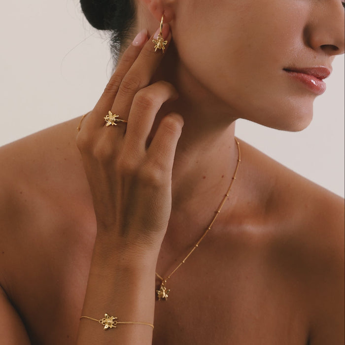 Woman wearing gold jewelry including earrings, a necklace, and bracelet against a neutral background