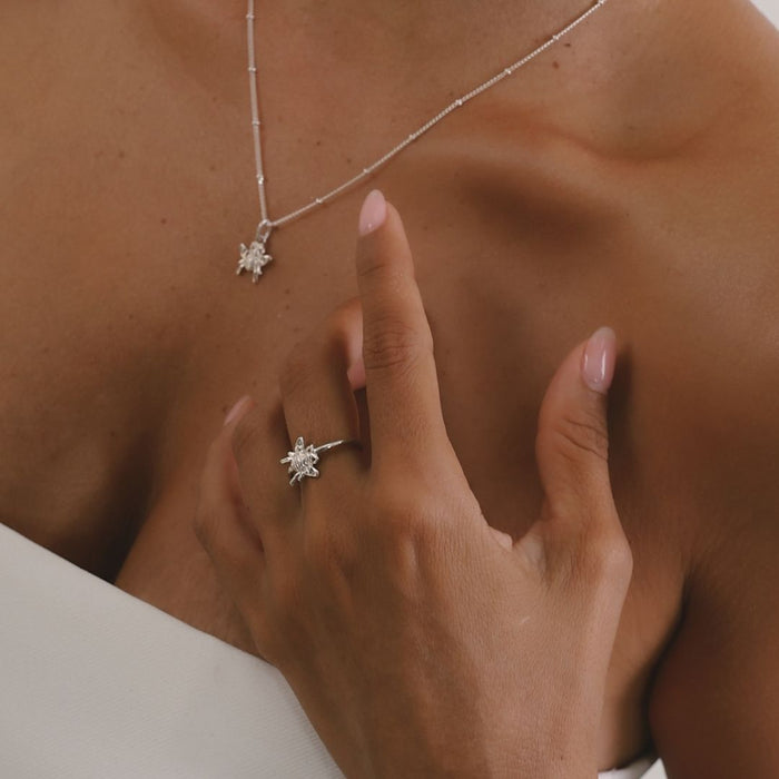 Close-up of a necklace and ring with a hand touching the chest, on a neutral background.