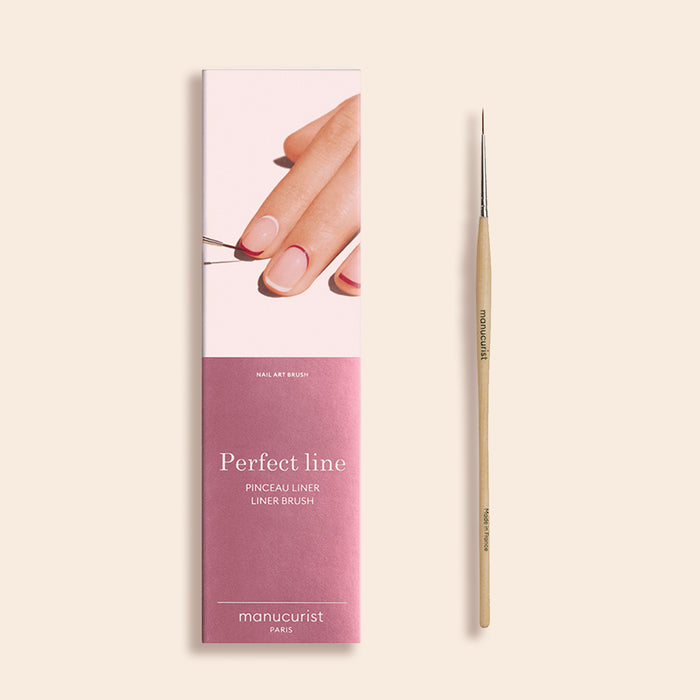The Manucurist Liner Brush, a thin nail art tool with a wooden handle and vegan synthetic bristles, is shown next to its pink and white packaging featuring a hand painting nail art and the text "Perfect line liner brush" and "manucurist PARIS.