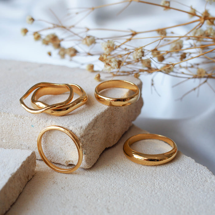 A display of gold rings with various designs, arranged on beige stone with dried flowers.