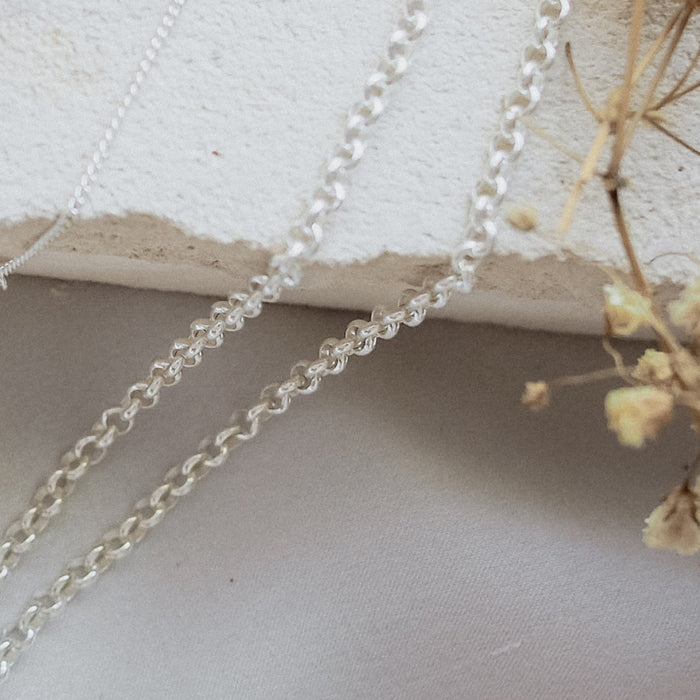 A silver chain bracelet with an adjustable clasp and circular charm, displayed on beige stone with dried flowers.