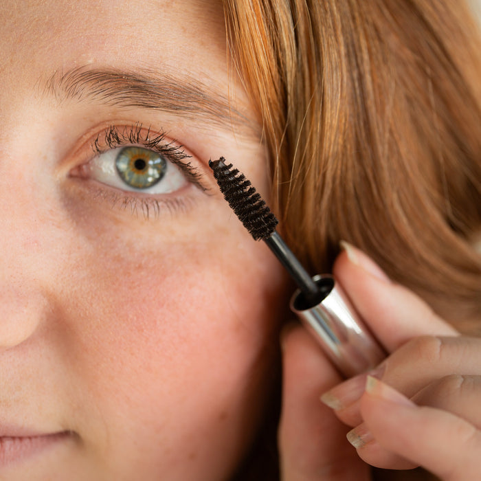 Close-up of a woman applying black mascara with a spoolie wand to her upper lashes, holding the silver and black tube in her hand.