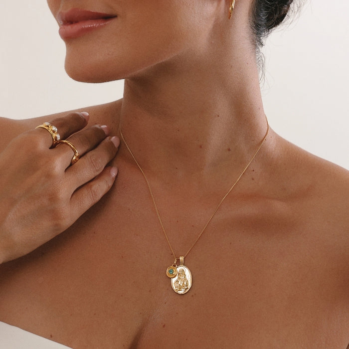 Close-up of a woman wearing a gold necklace with a pendant, with a neutral background.