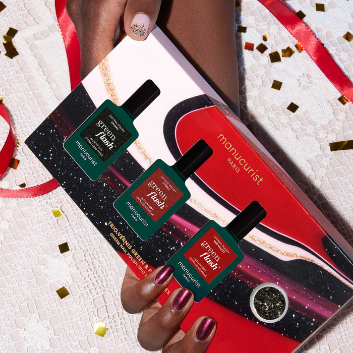 A glittery-manicured hand holds the Manucurist-test Midnight Glam Trio + Glitter Set gift box, displaying three green polishes with biodegradable glitter, on a lace table decorated with gold confetti.