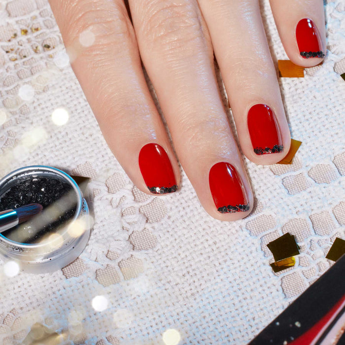 A hand with red polish and black glitter tips from Manucurist-test’s Midnight Glam Trio + Glitter Set rests on lace, with a brush and biodegradable glitter nearby and gold confetti scattered around.