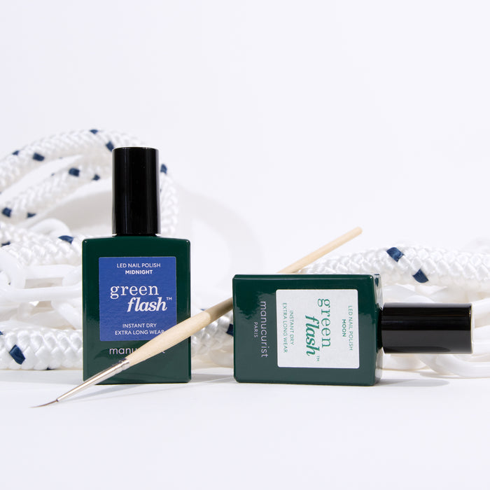 Two bottles of Manucurist Midnight Swim nail polish are shown with a wooden cuticle stick, in front of a coiled white rope with navy accents on a white background—ideal for creative nail art.