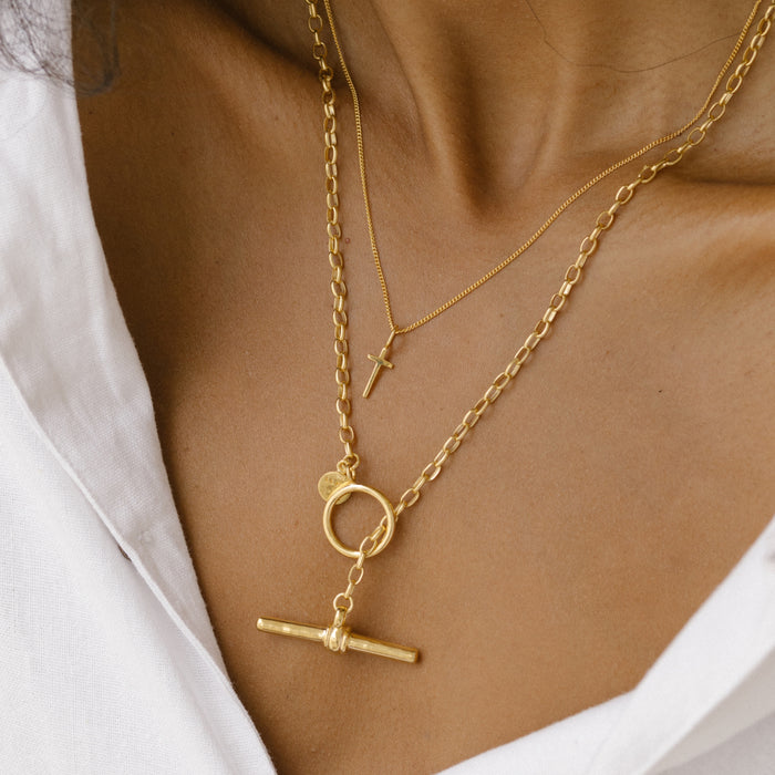 A close-up of layered gold necklaces, including one with a small cross pendant and another with a circular and bar design, worn with a white shirt.