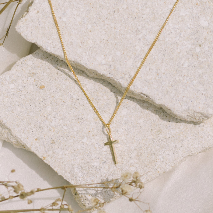A gold necklace with a small cross pendant, laying on textured stones.