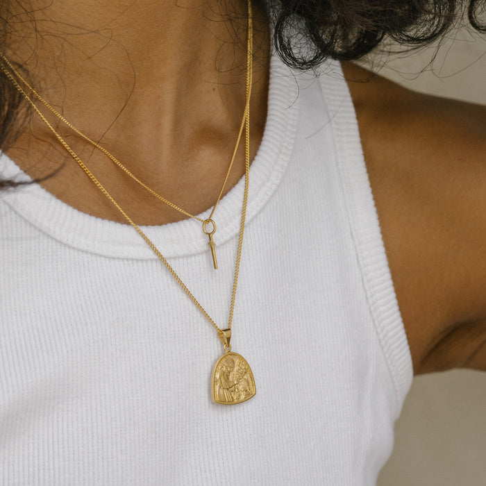 A close-up of layered gold necklaces, including one with a small key pendant and another with a larger gold medallion, worn with a white ribbed tank top.