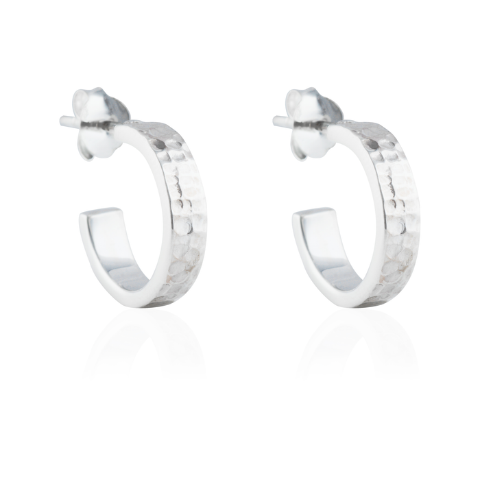 Close-up of a pair of hammered silver hoop earrings with post backings on a reflective white background.