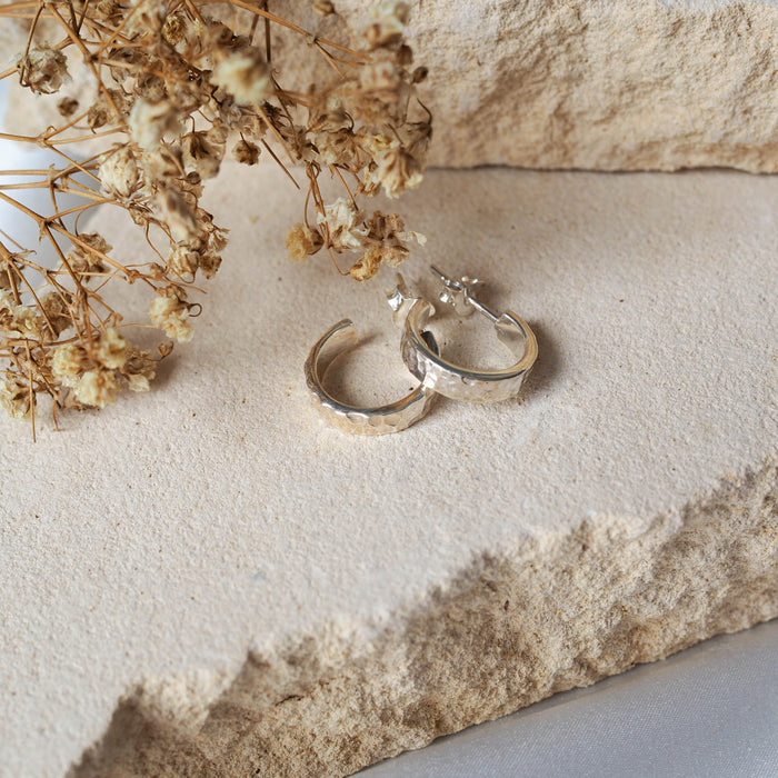 A pair of hammered silver hoop earrings displayed on textured stone beside dried flowers.