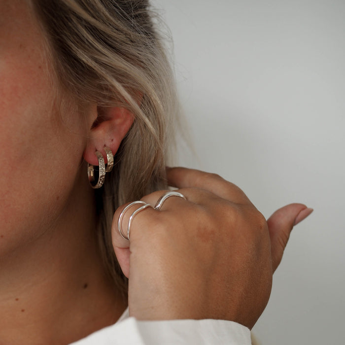A person wearing two hammered silver hoop earrings in one ear, with silver rings on their fingers.