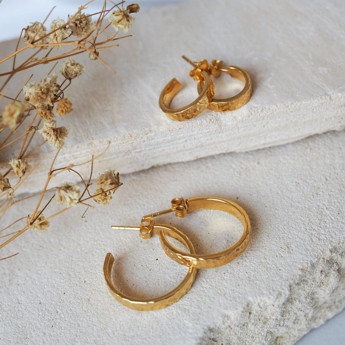 Two pairs of hammered gold hoop earrings displayed on textured stone beside dried flowers.
