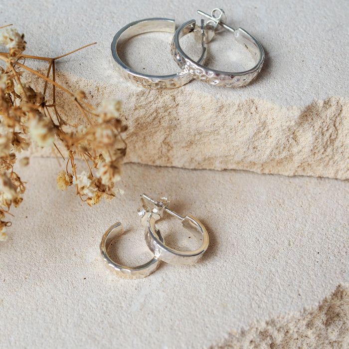 Two pairs of hammered silver hoop earrings displayed on textured stone beside dried flowers.