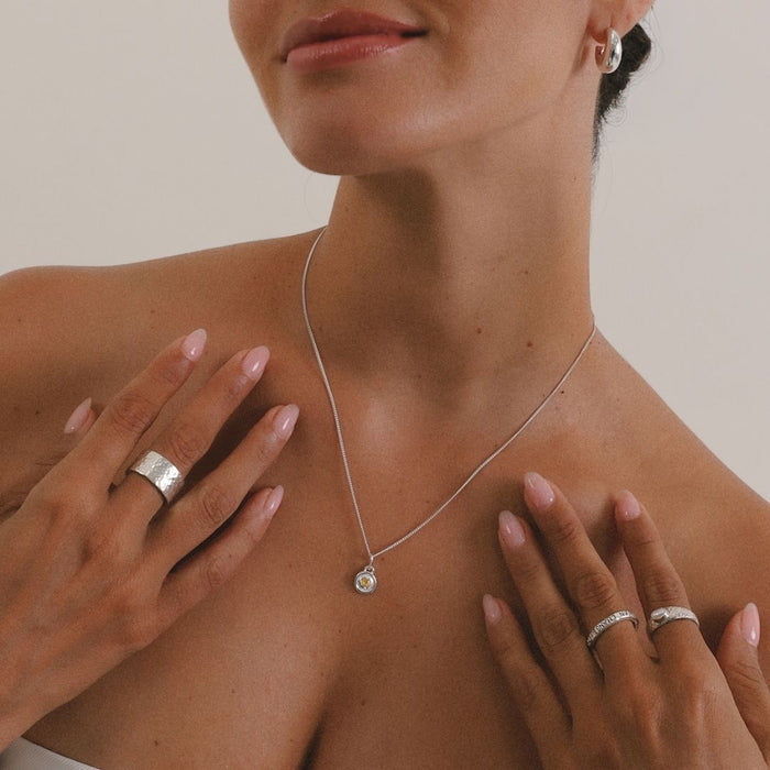 Woman wearing a silver necklace with a pendant, with her hands gently touching her shoulders.