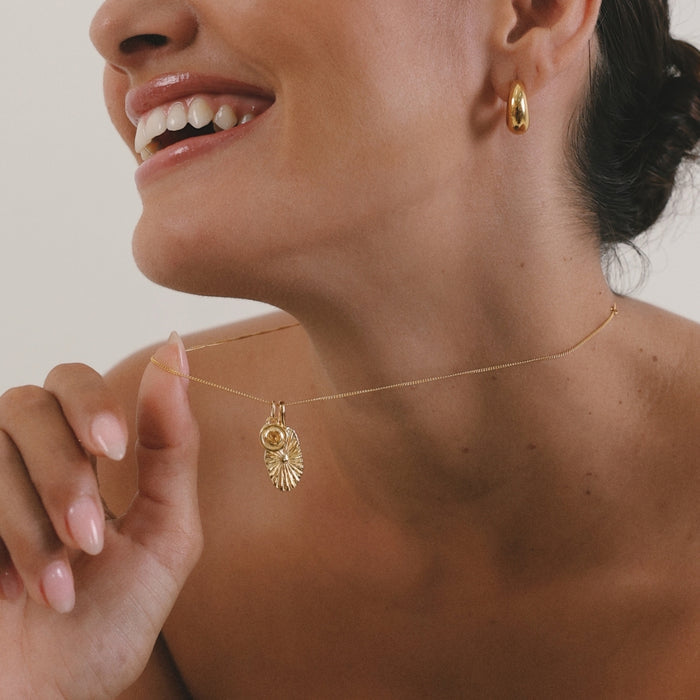 Close-up of a woman wearing a gold necklace with a pendant, against a neutral background.