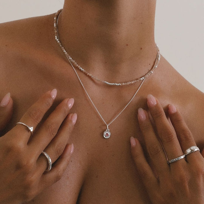 Close-up of a person wearing a silver necklace with a pendant, surrounded by hands with rings.