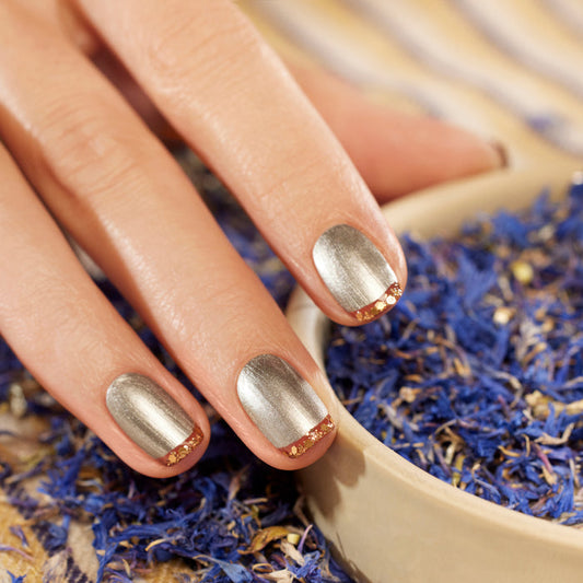 A hand with metallic silver Manucurist Oriental Vibe nail polish and gold-tipped accents rests beside a bowl of dried purple and blue flower petals.
